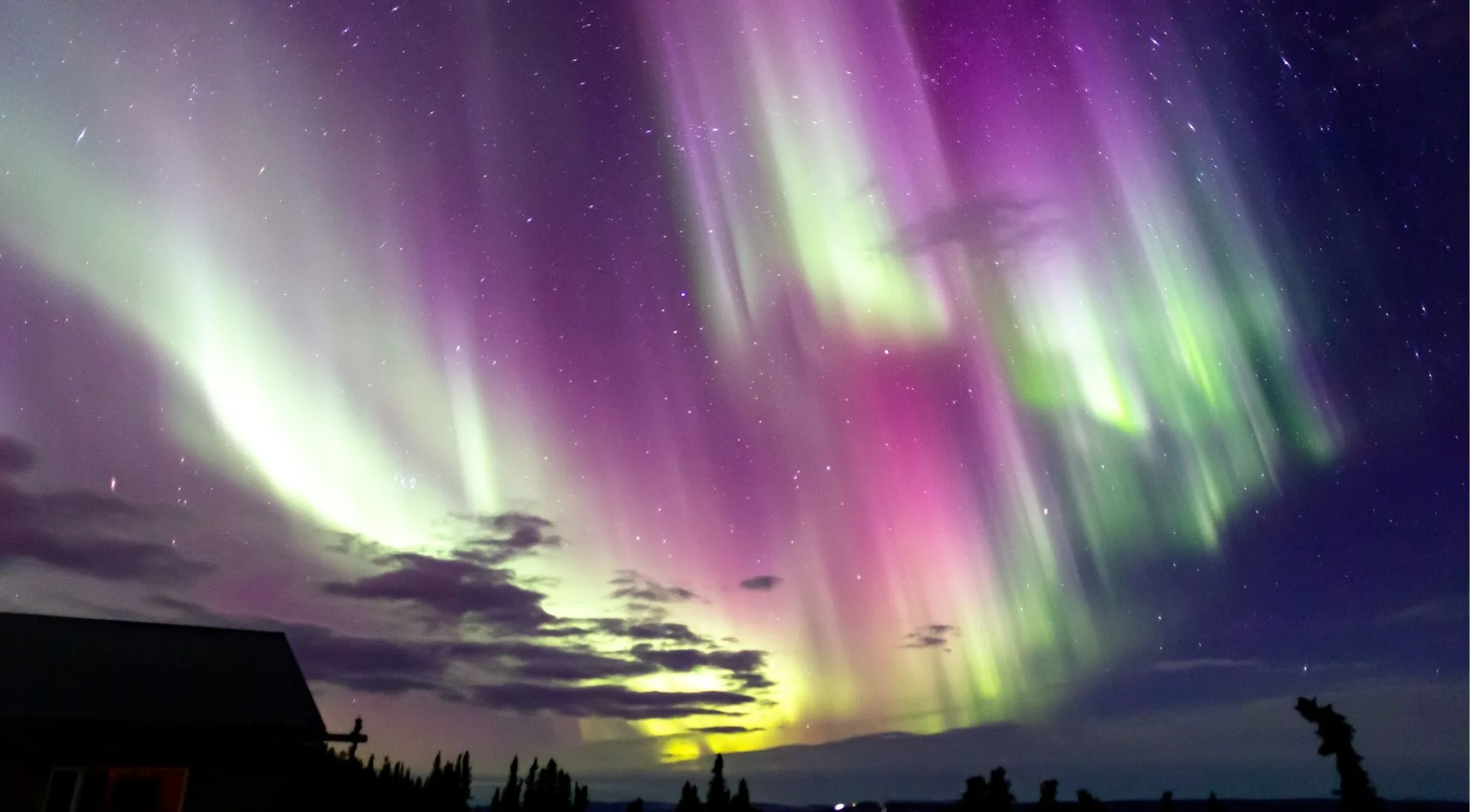 Pink and green aurora curtains above a dark roofline with thin clouds drifting across