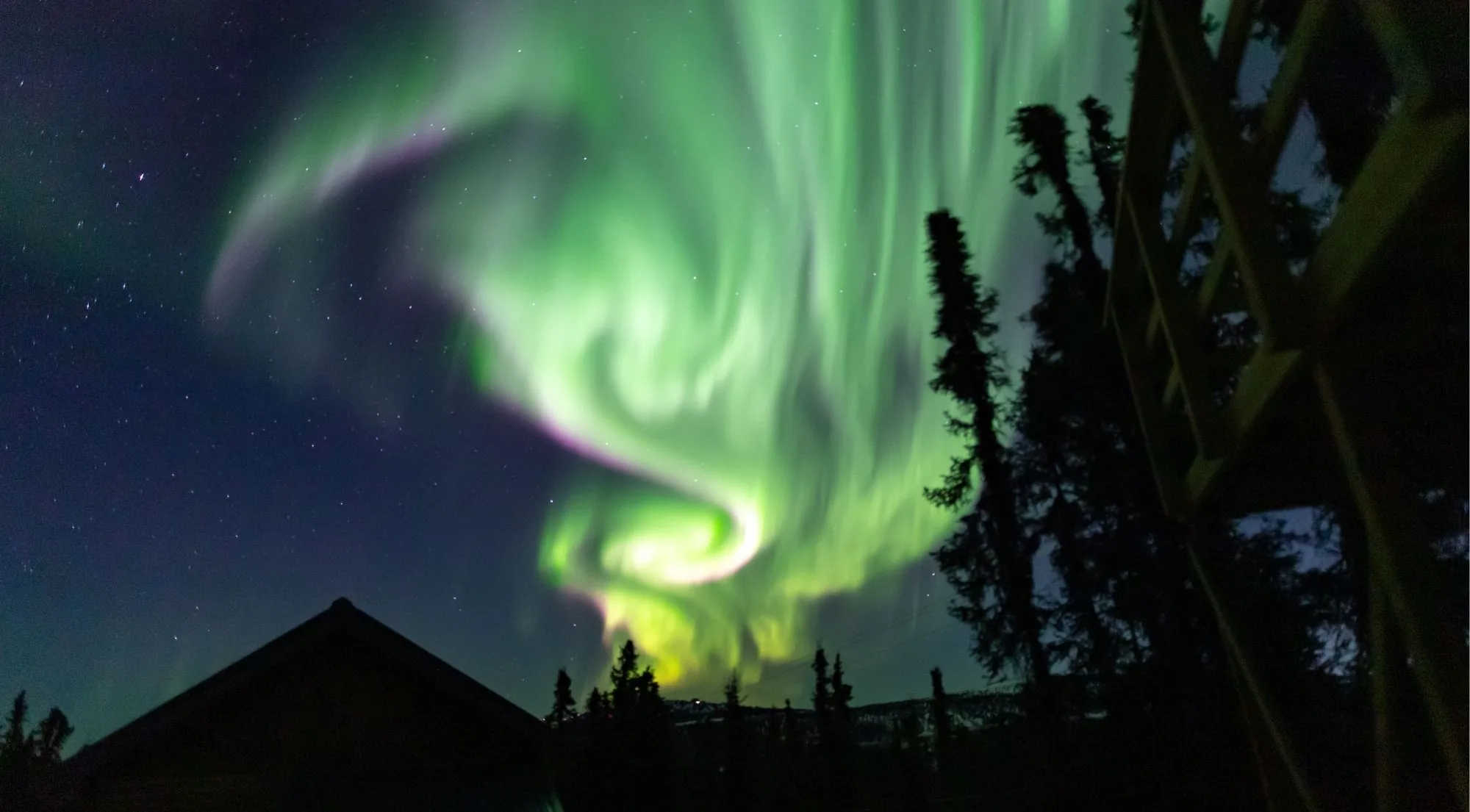 A swirling green aurora above a cabin roof and tall spruce trees