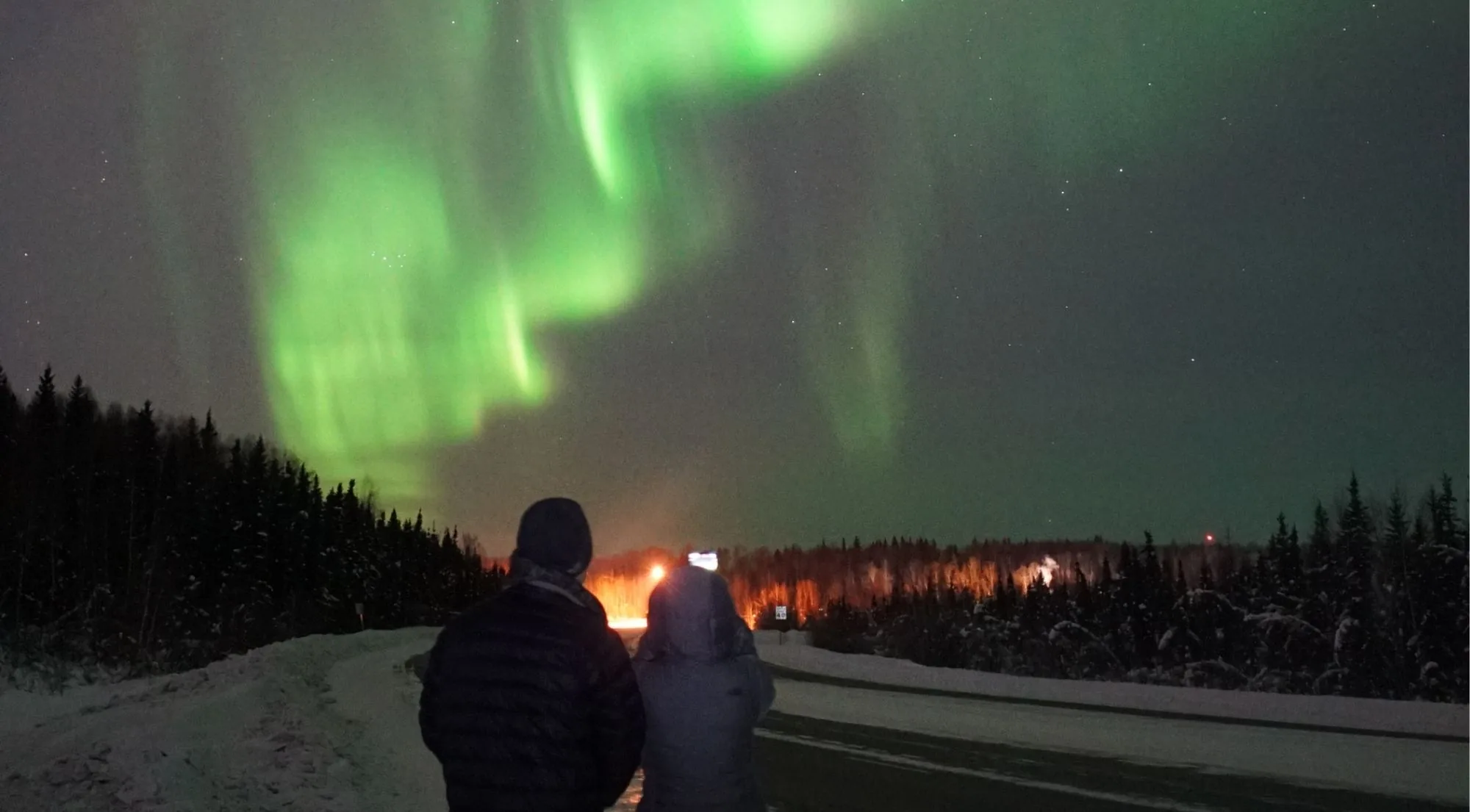 Two people in winter coats watching a green aurora over a snowy forest edge at night