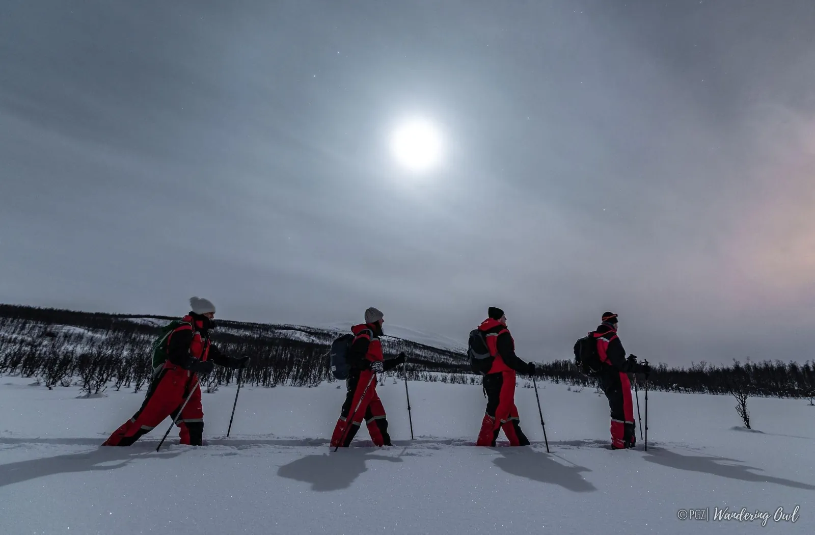 Four snowshoers in red thermal suits walking through deep snow under a bright moon