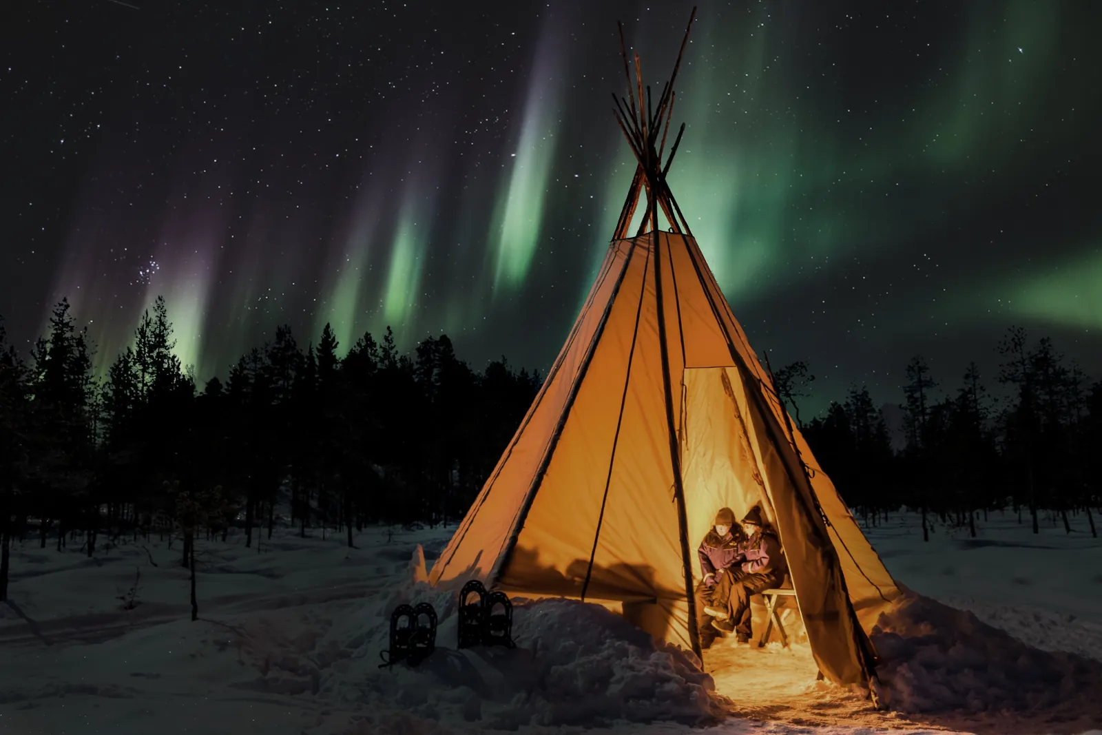 Two guests sit inside a lit tipi tent under green aurora, with snowshoes outside