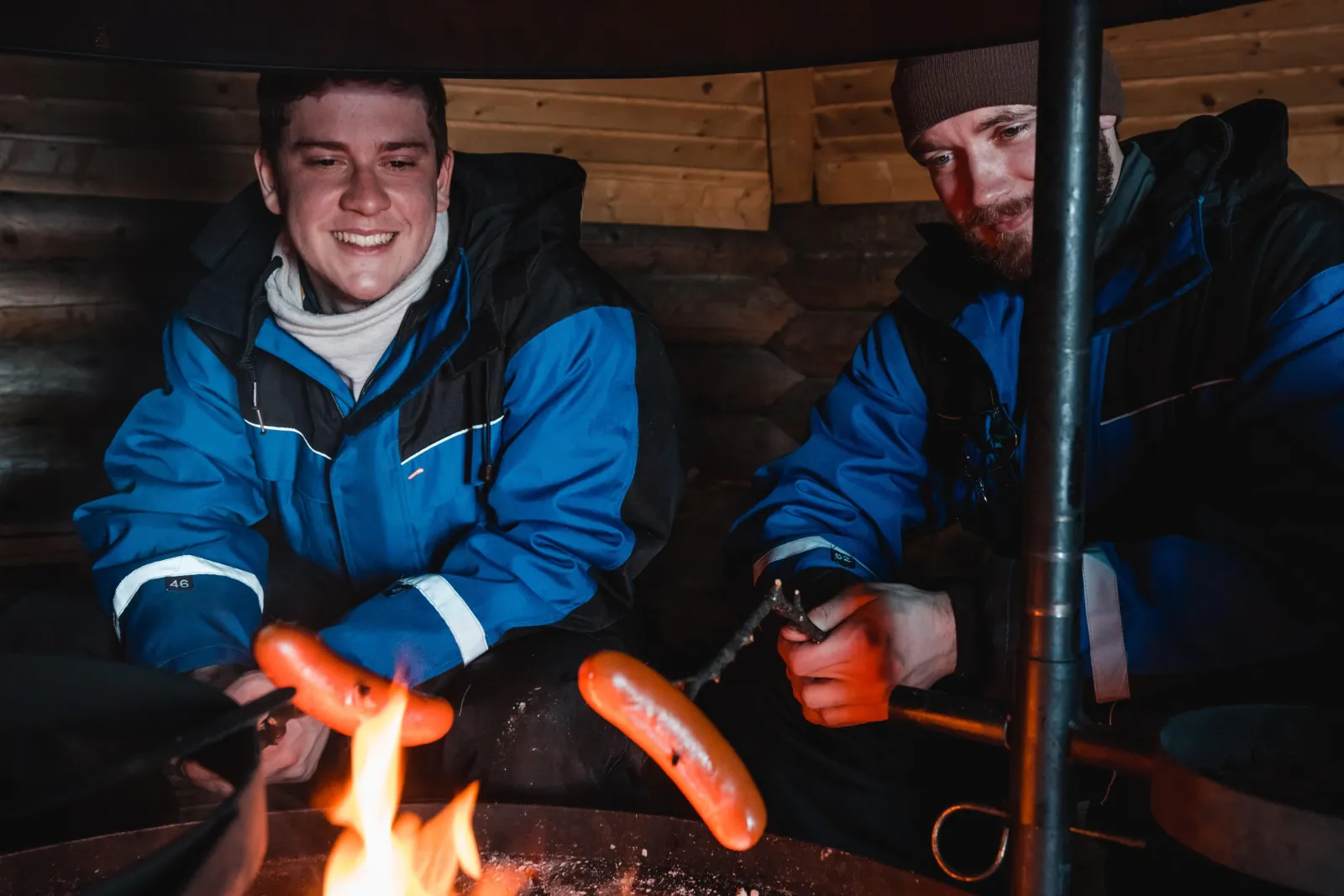 Two guests in blue snowsuits roast sausages over a fire inside a wooden hut