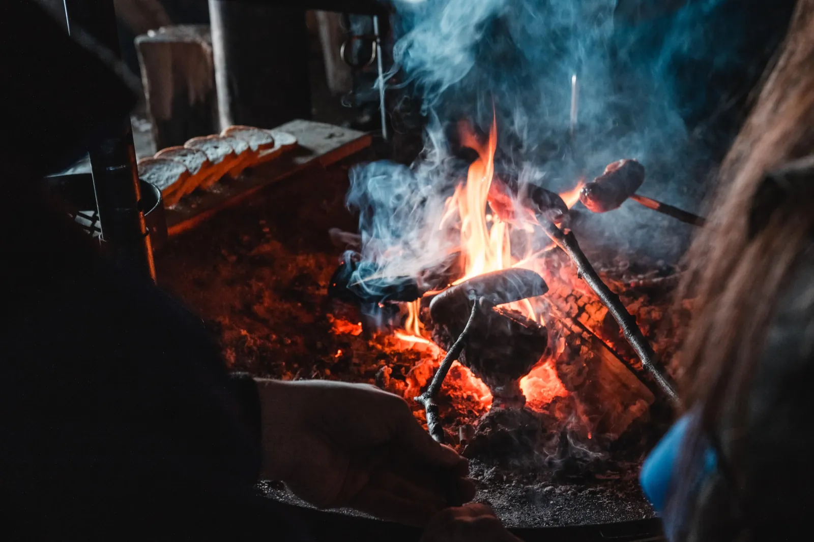 Sausages on sticks over a smoky fire, with sliced bread on a board nearby