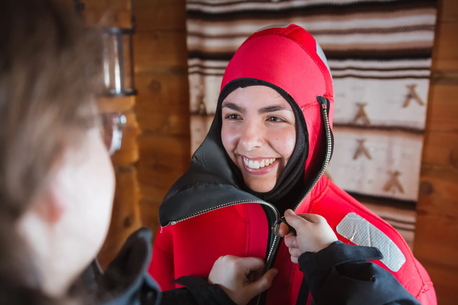 A guest being zipped into a red floating suit inside a wooden cabin