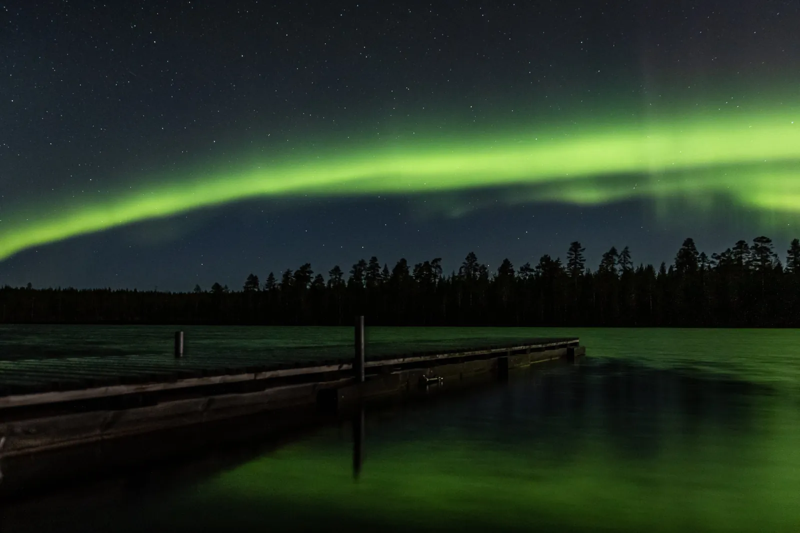 A green aurora arc over a dark lake with a wooden jetty and pine treeline