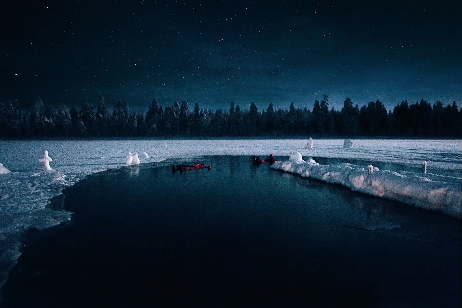 Guests in floating suits in open water cut into a snowy frozen lake at night