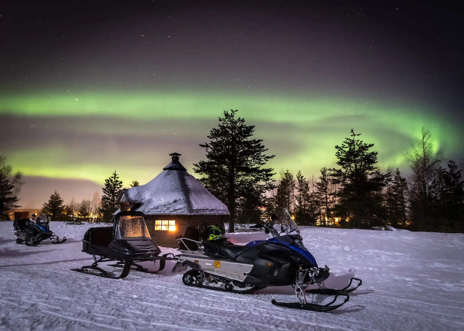 A snowmobile with a covered passenger sledge beside a lit wooden hut under green aurora