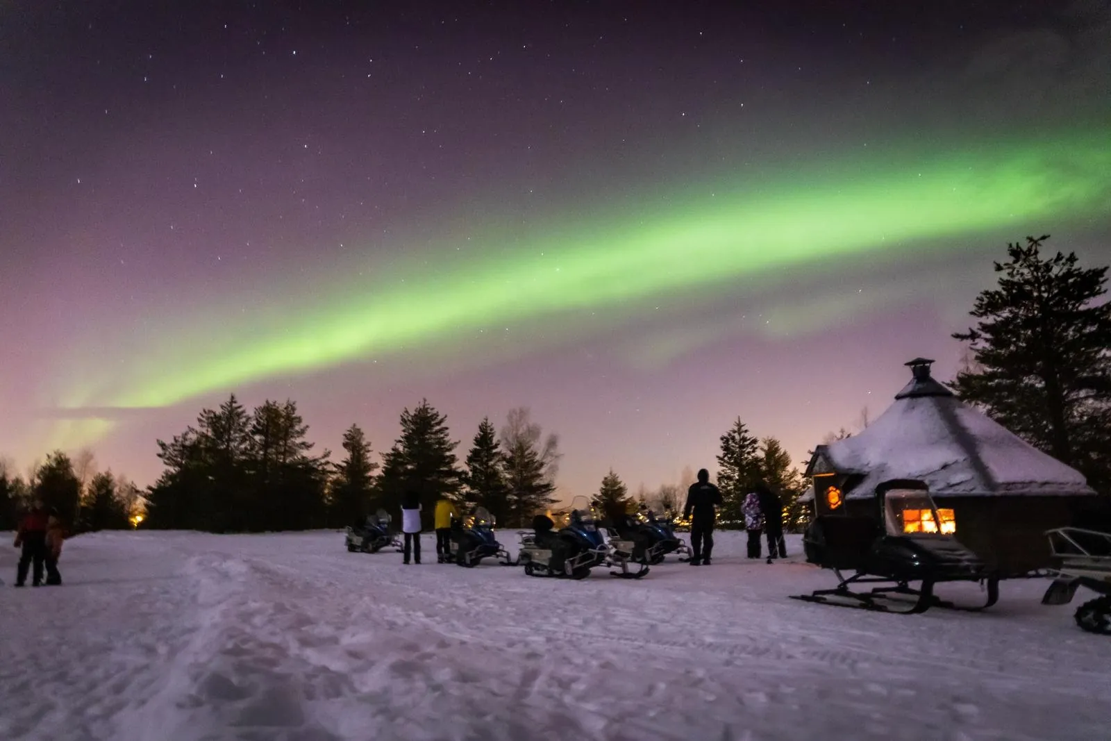 Guests stand by parked snowmobiles and a lit hut as a green aurora crosses the sky
