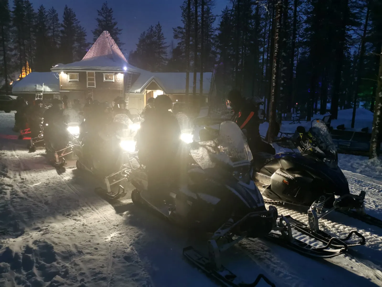 Guests on snowmobiles with headlights on, lined up outside a snow-covered lodge at night