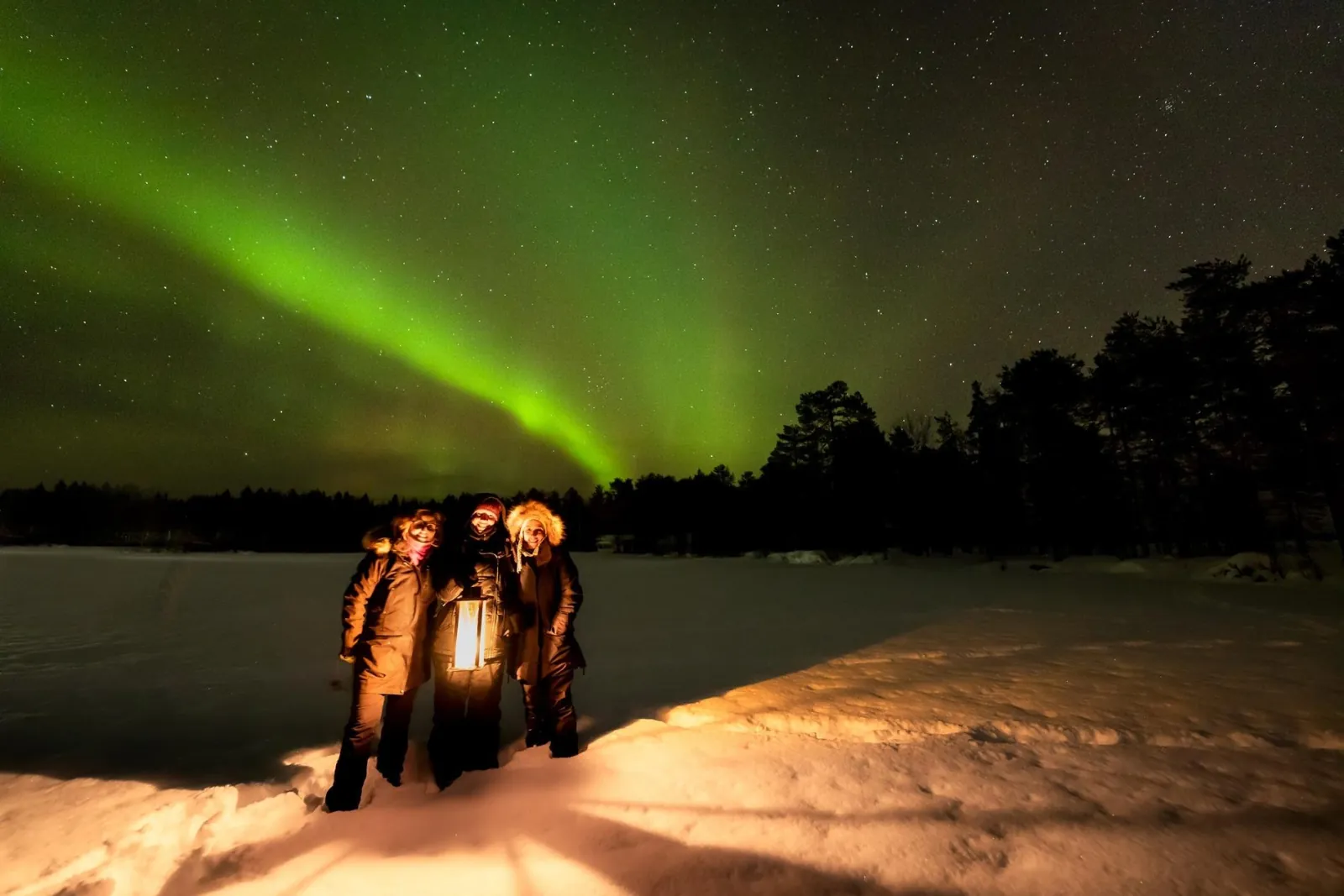 Three guests holding a lantern on a snowy frozen lake under a green aurora arc