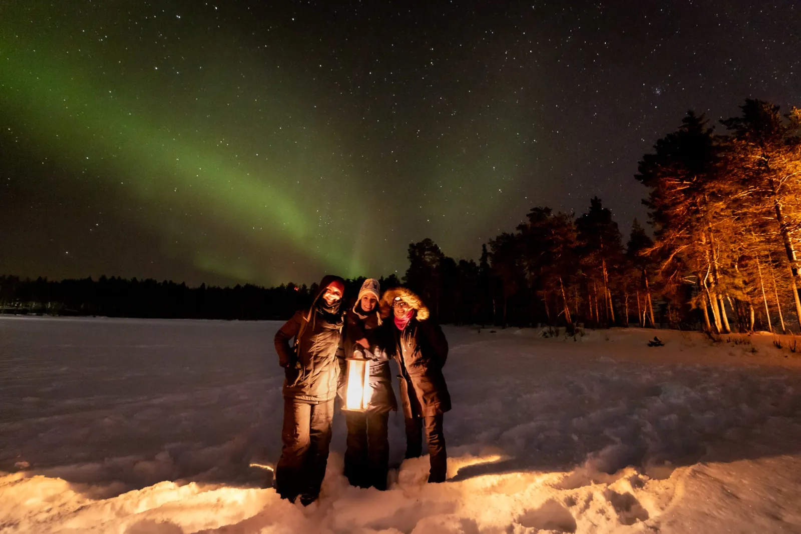 Three guests with a lantern on snow under a faint green aurora, trees lit orange