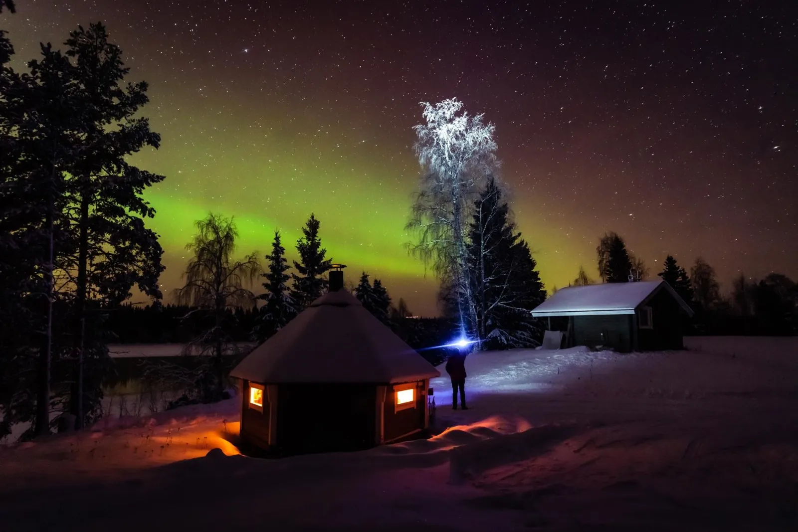 A lit wooden hut and a snowy cabin under a green aurora and starry sky