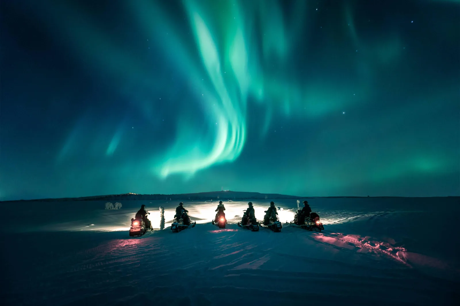 Six snowmobiles with headlights on face a large green aurora over open snow