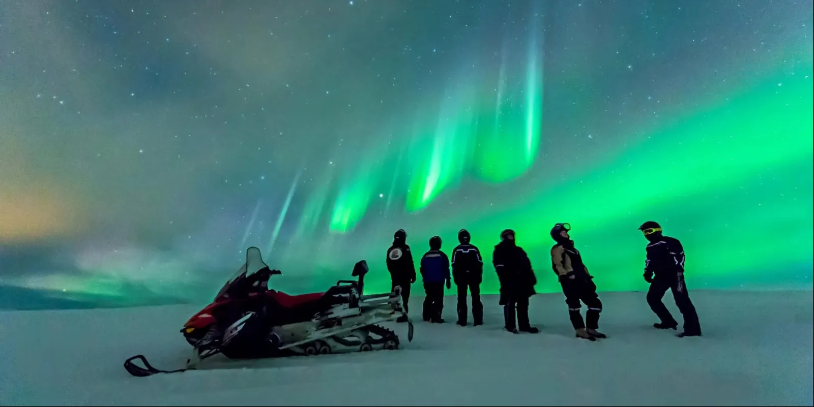 Guests in snowsuits and helmets look up at green aurora beside a red snowmobile