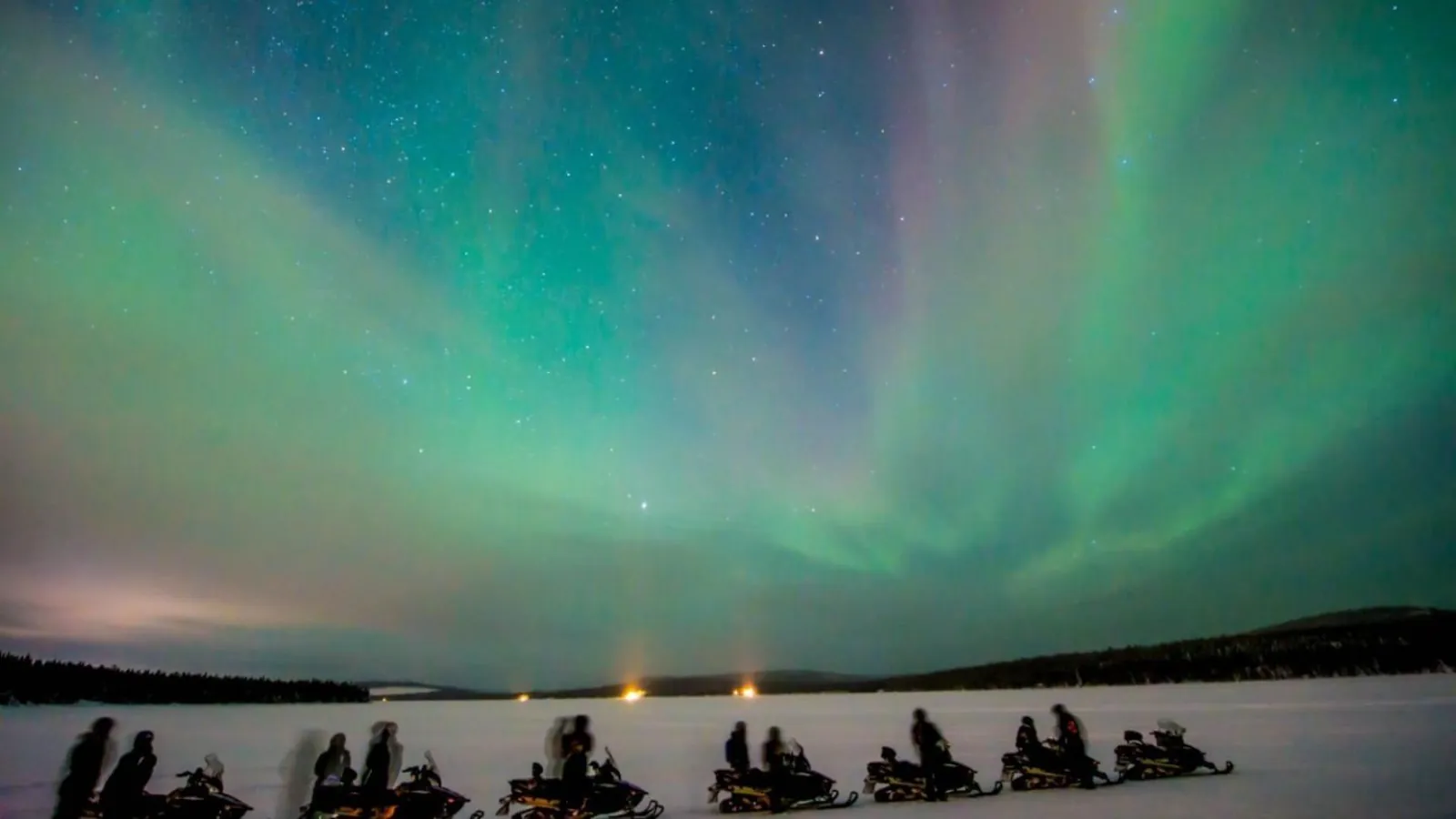 A row of snowmobiles and riders on a frozen open plain under green aurora