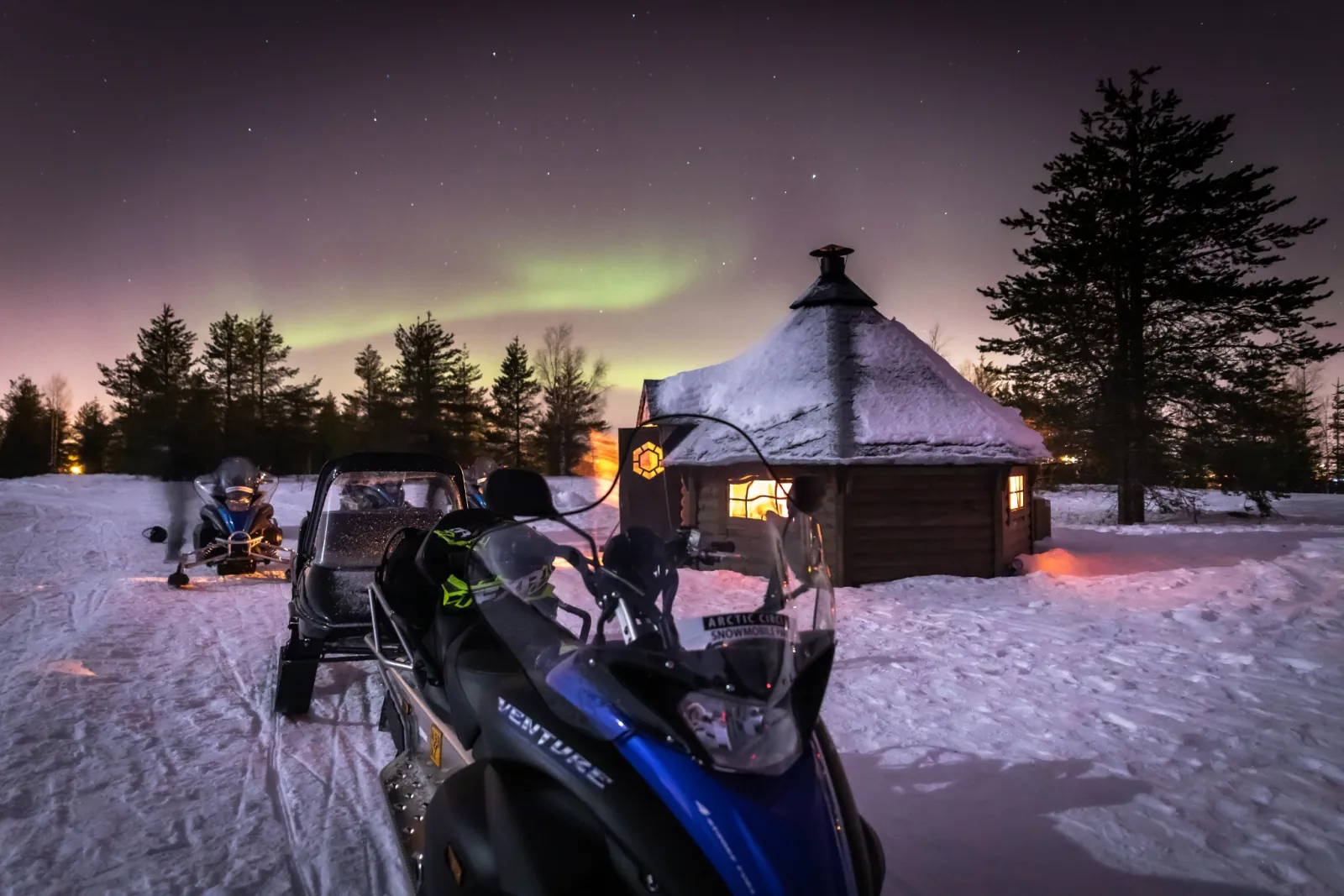 Parked snowmobiles beside a lit wooden hut, with a faint green aurora above the trees