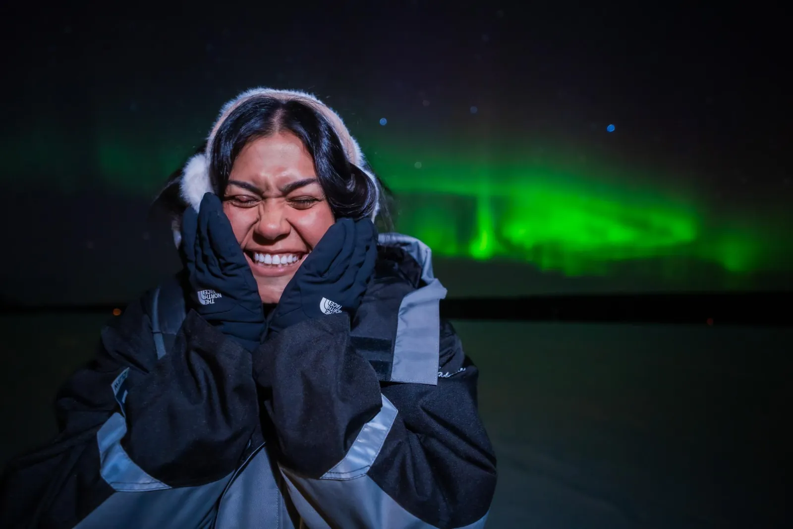 A guest in black mittens holding their cheeks under a low green aurora band