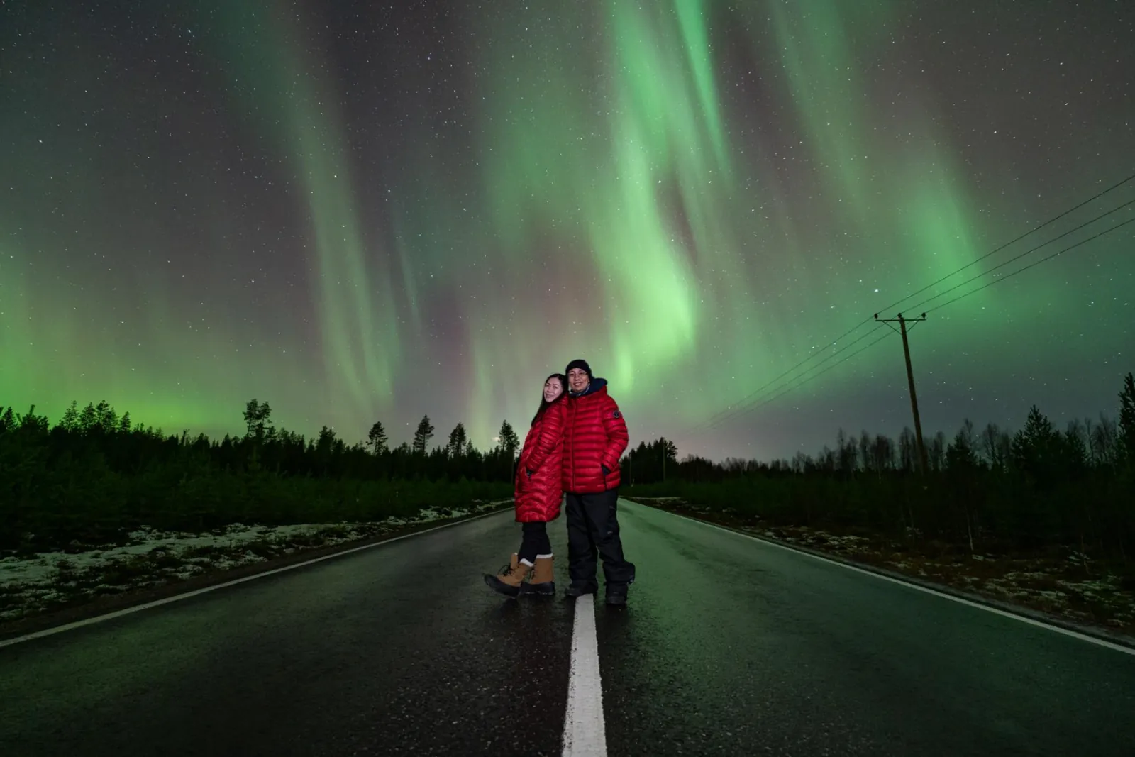 Two guests in red down jackets on a wet forest road under green aurora curtains