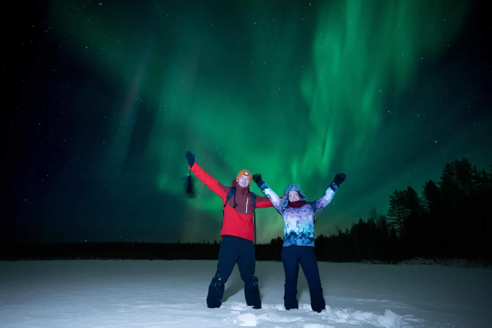 Two guests with raised arms on snow under a green aurora above dark spruce trees