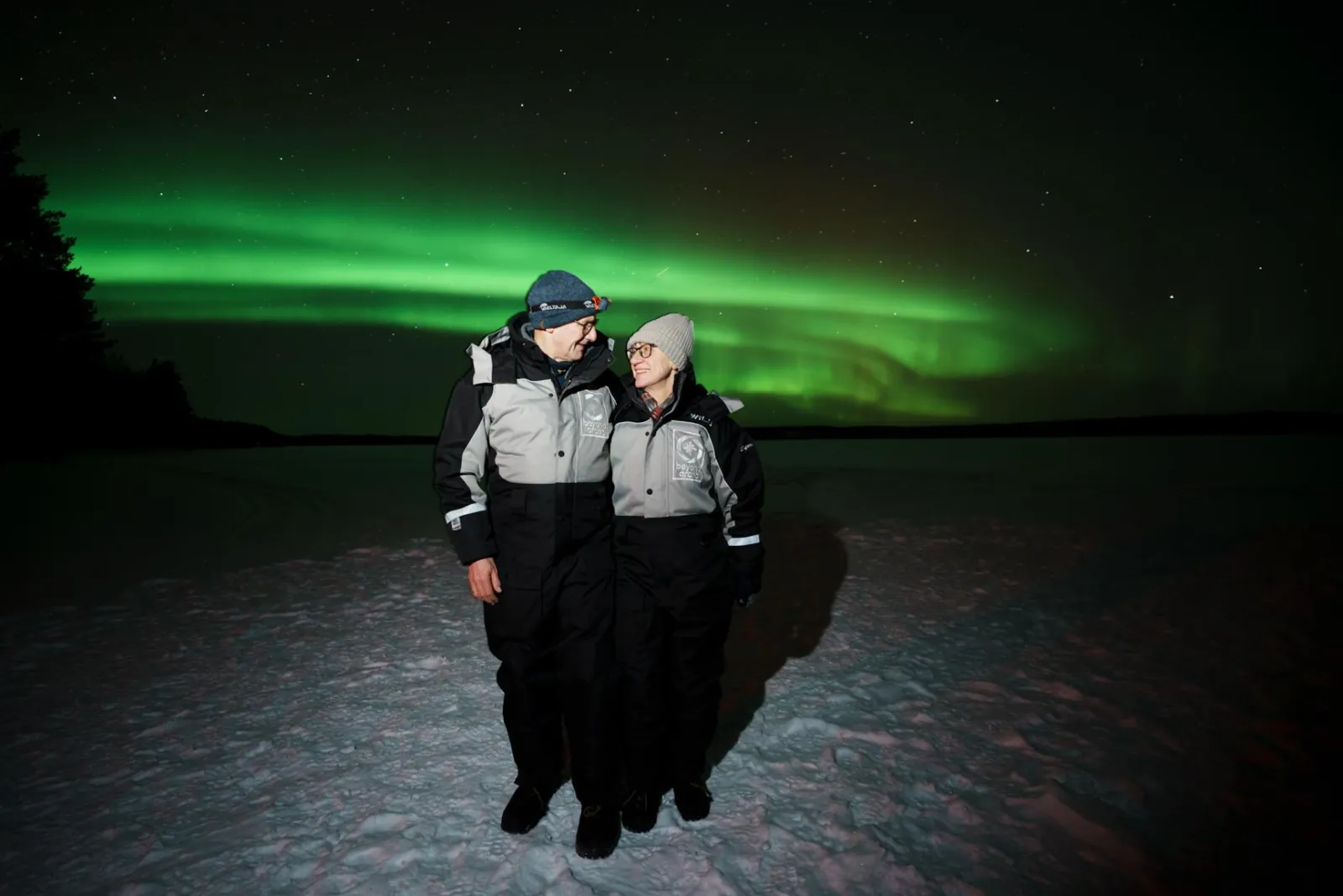 Two guests in grey and black overalls on a snowy lake under a green aurora arc