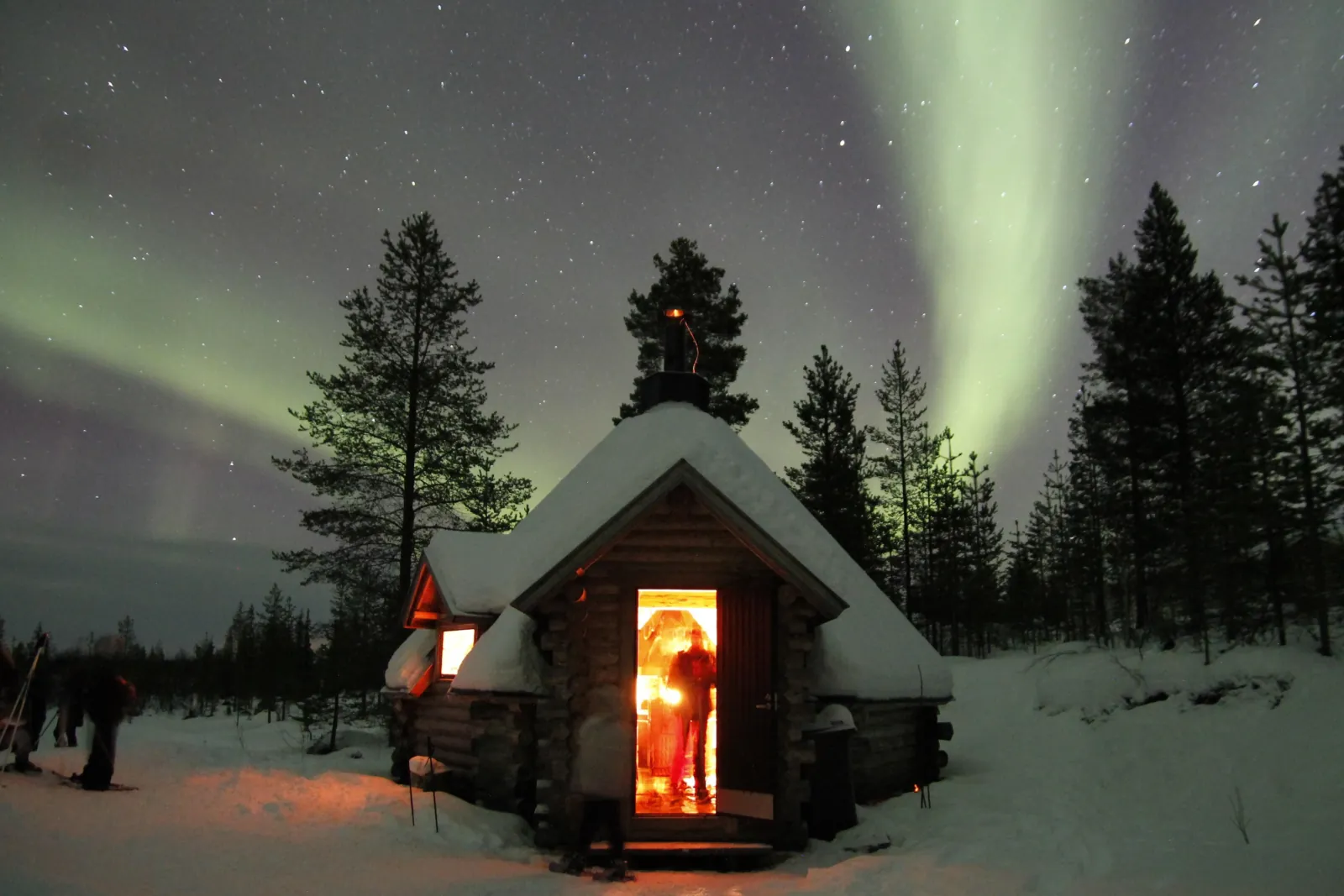 A lit log hut with an open door under a starry sky and pale green aurora