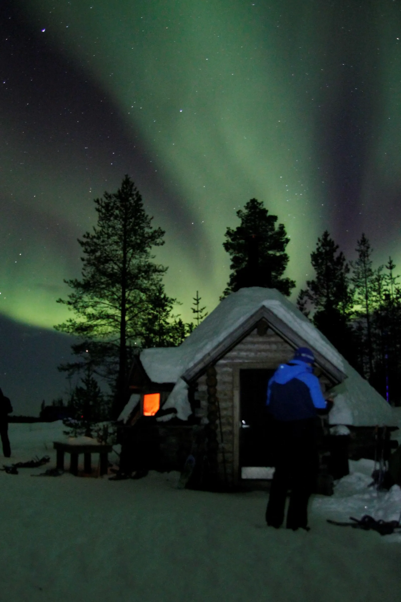 A guest in a blue jacket outside a snowy log hut under green aurora