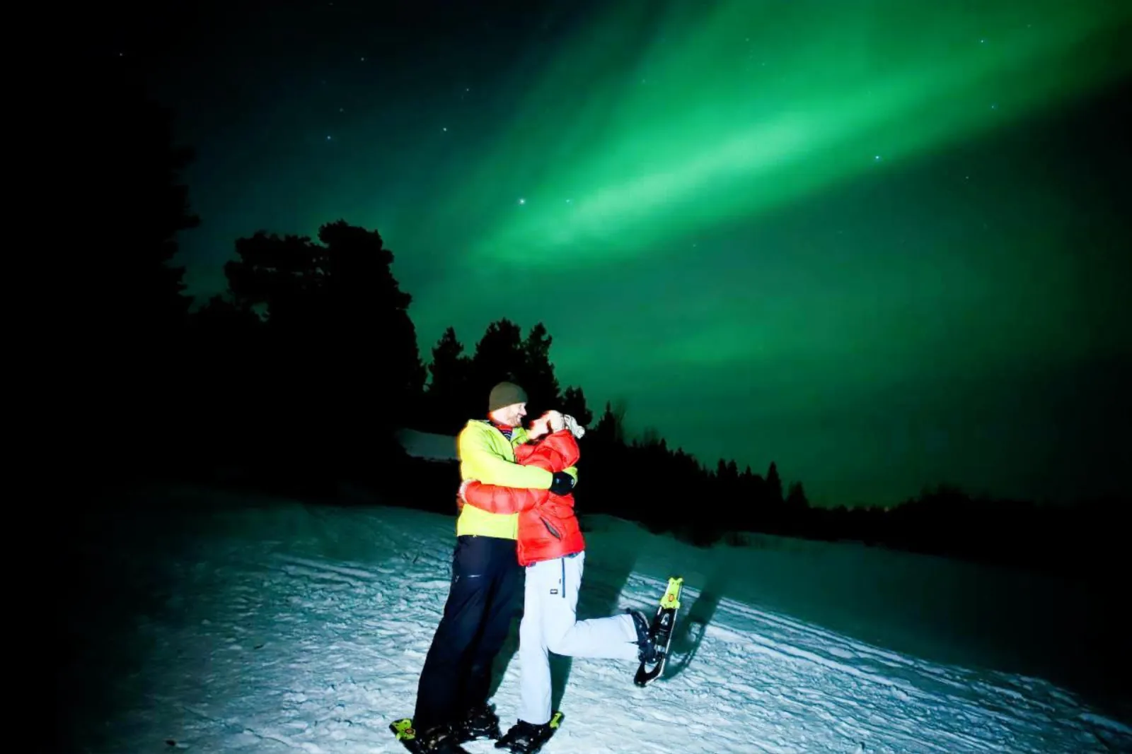 Two guests on snowshoes hugging under a broad green aurora band at night