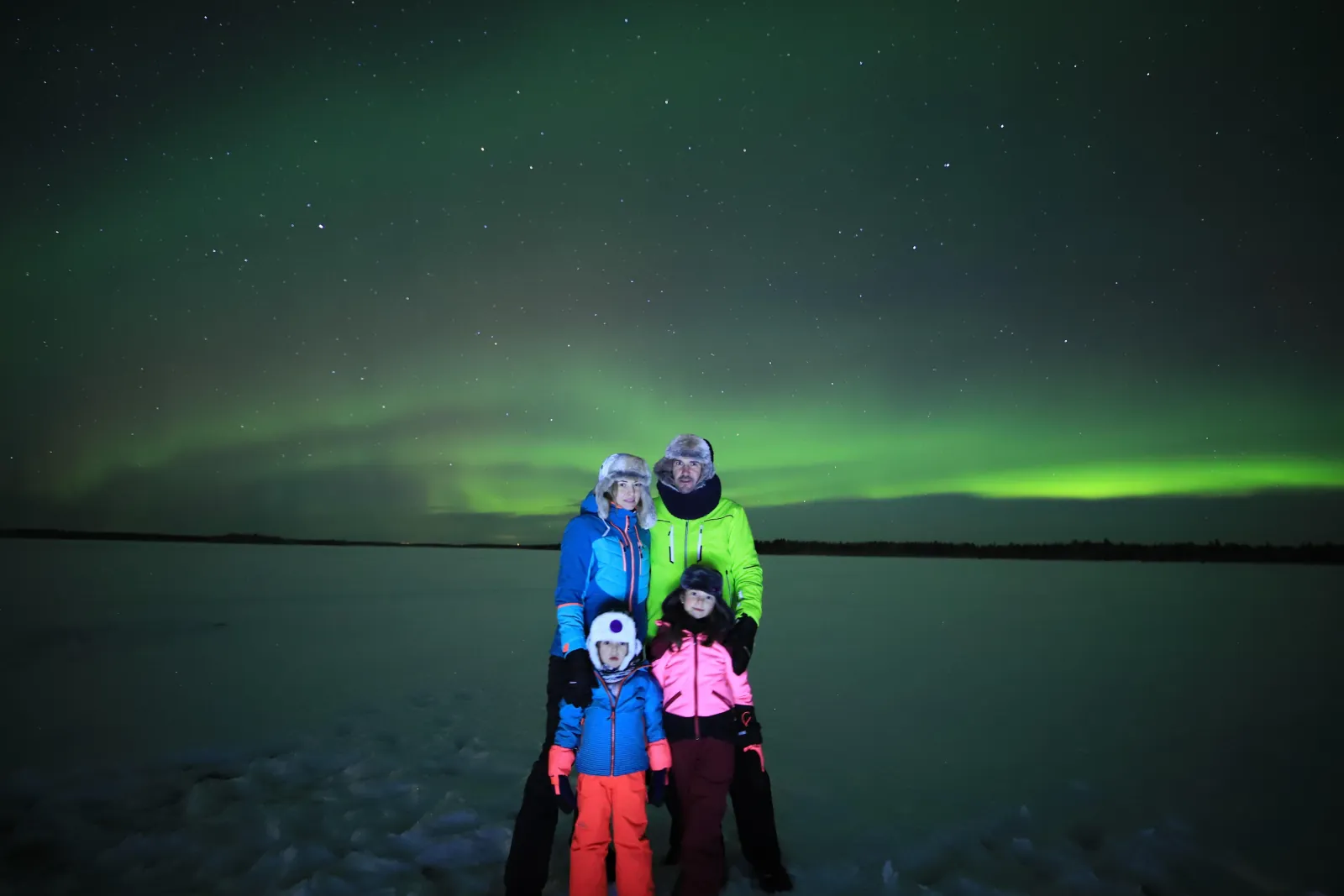 A family of four in colorful snowsuits on a frozen lake under a low green aurora