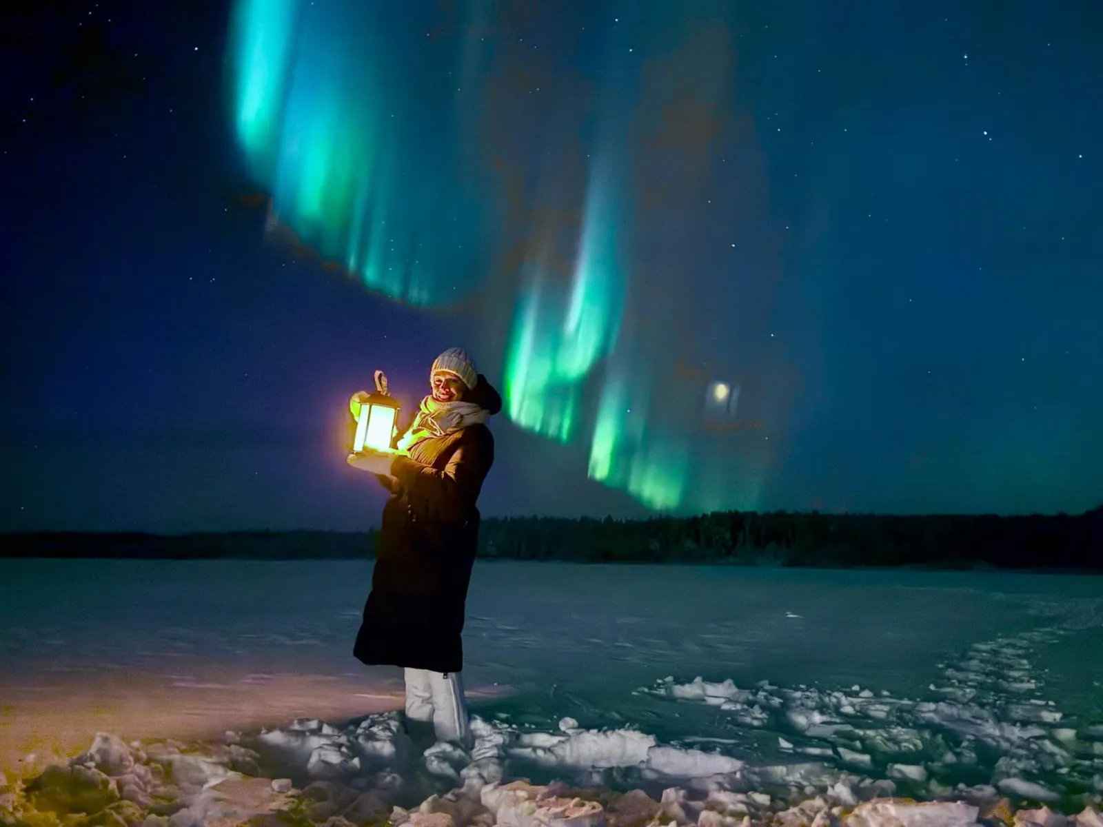 A guest holding a glowing lantern on a frozen lake under green aurora curtains