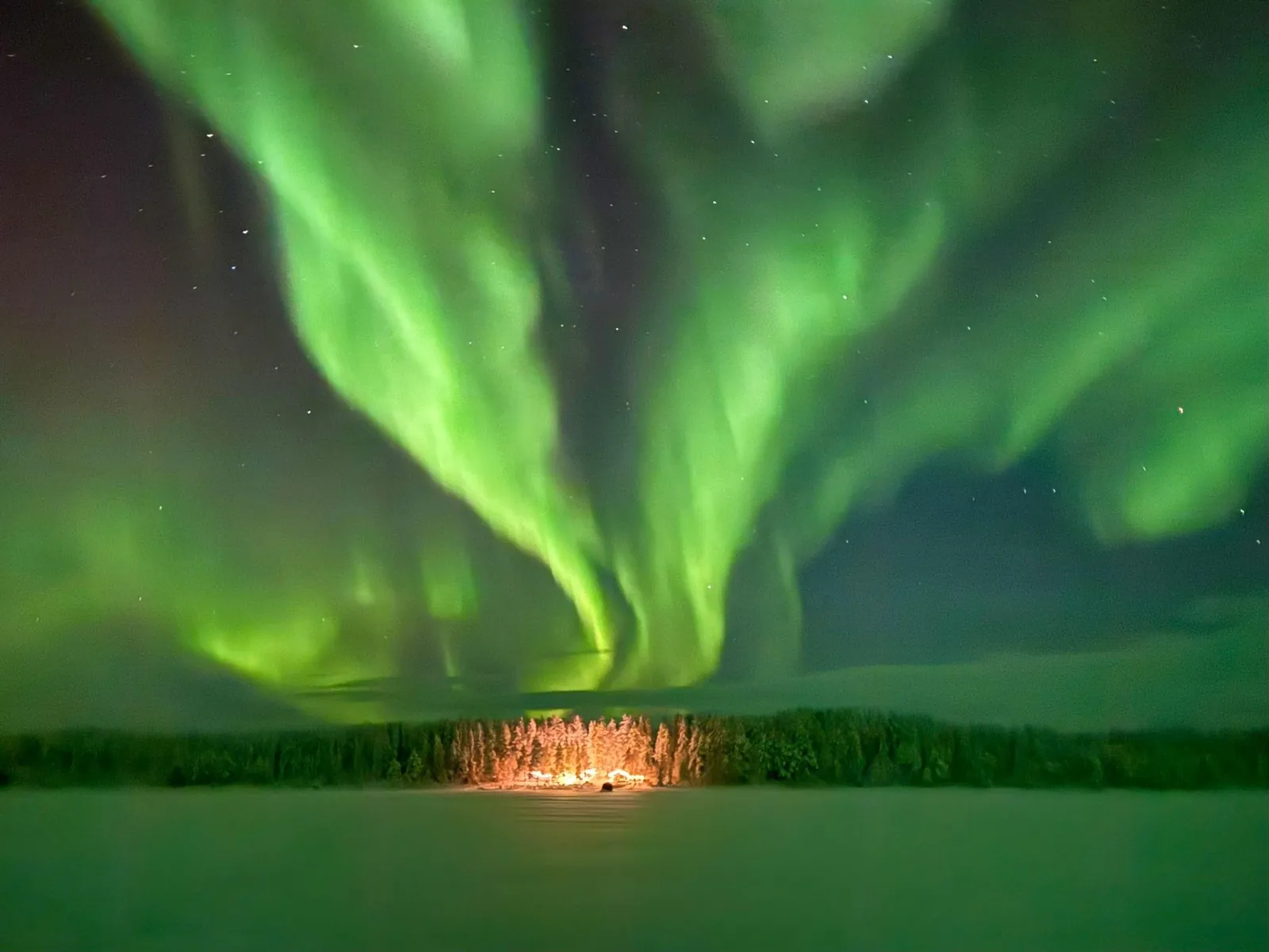 Bright green aurora swirling above a frozen lake and a lit cabin among snowy trees