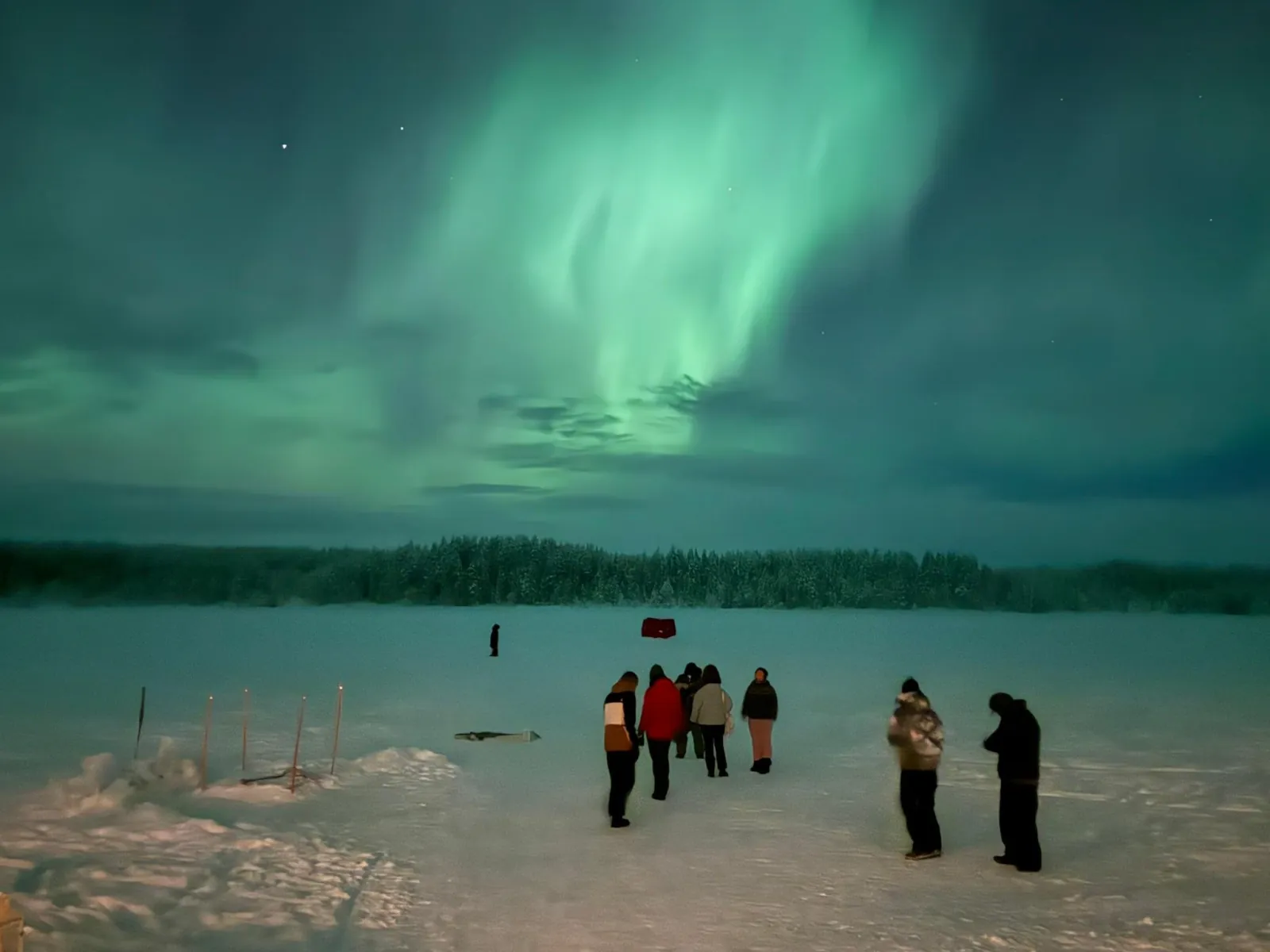 A small group standing on a snowy frozen lake watching a green aurora over forest