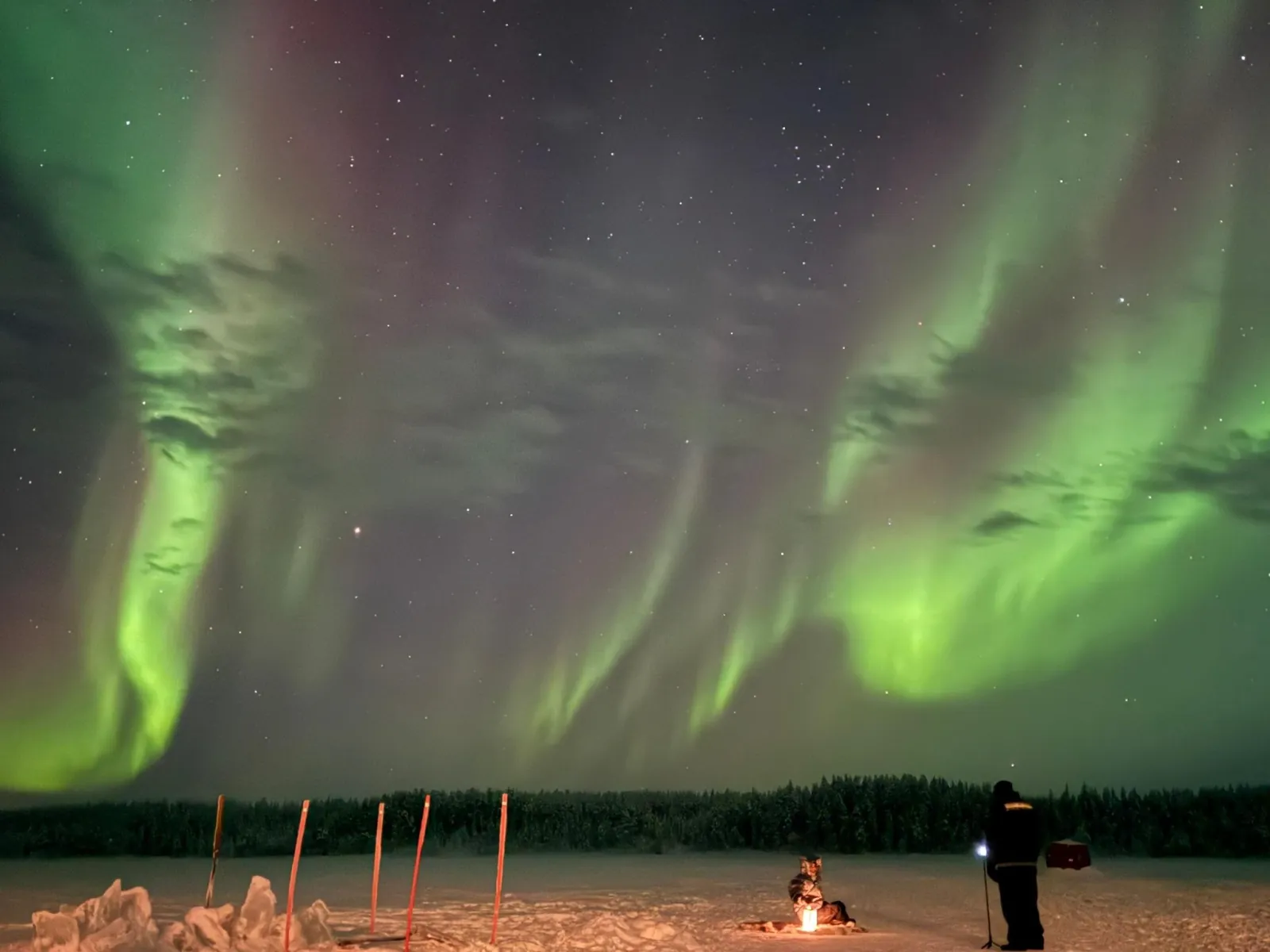 Green and faint red aurora over a frozen lake with marker poles and a lantern