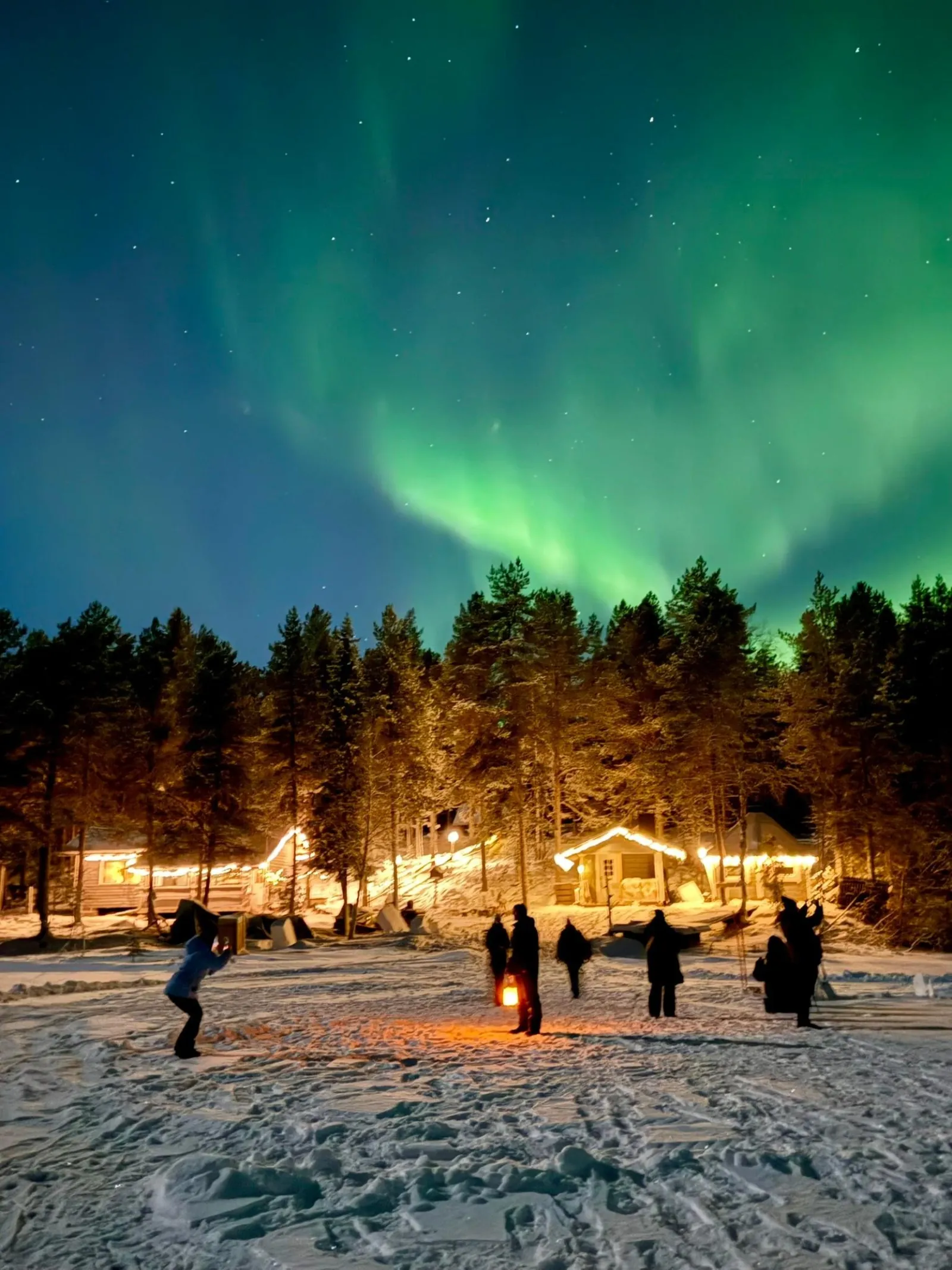 Guests with a lantern on a snowy lake below green aurora and lit wooden cabins