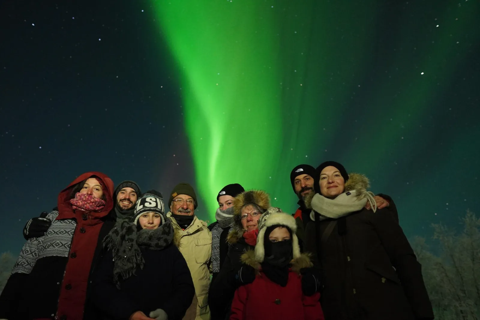 A family group in hats and scarves standing under a tall green aurora band