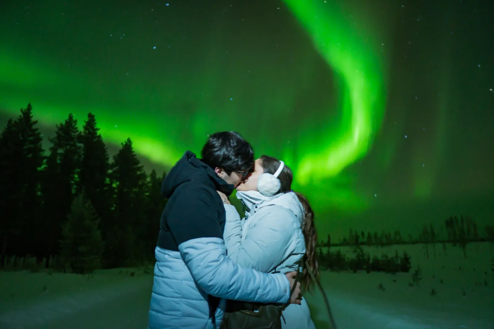 Two guests embracing on a snowy track beneath a curving green aurora and spruces