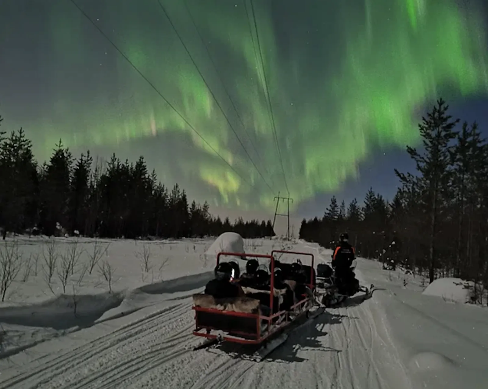 Guests in helmets ride a red open sledge behind a snowmobile under bright green aurora