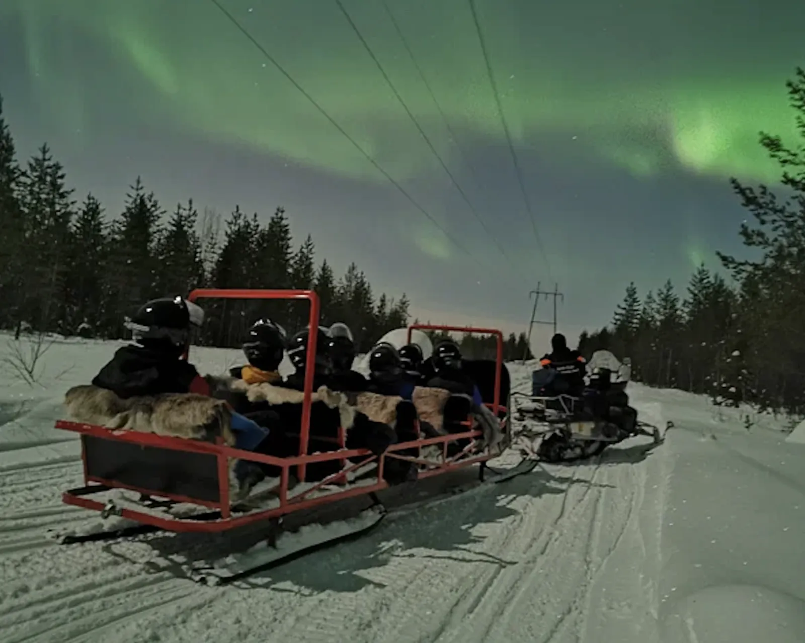 Helmeted guests sit on fur-covered seats in a red sledge towed by a snowmobile