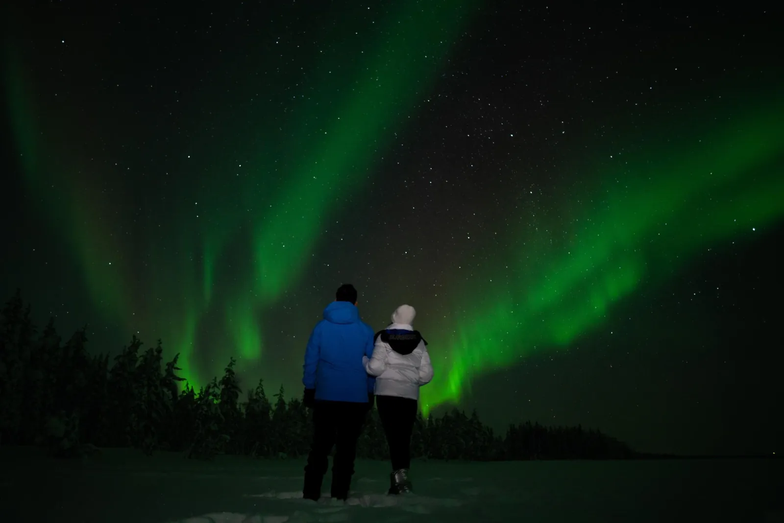 Two guests in winter jackets stand in snow watching green aurora above dark trees