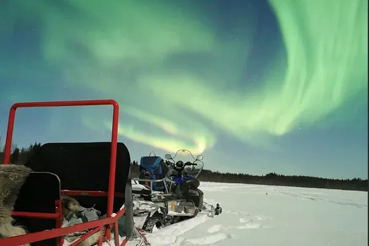 A red sledge pulled by a snowmobile, stopped on open snow under swirling green aurora
