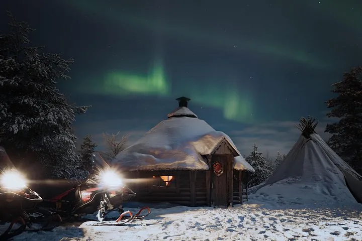 Snowmobiles with headlights on and a towed sledge beside a snowy hut and tipi under aurora
