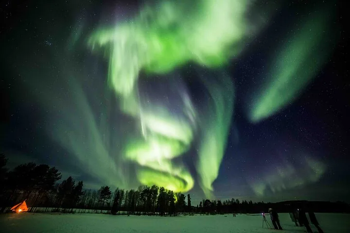 Bright green aurora swirls over snowy ground, a glowing tent and guests with tripods