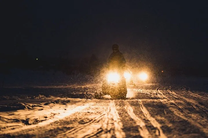 A snowmobile rider with headlights on drives through falling snow at night