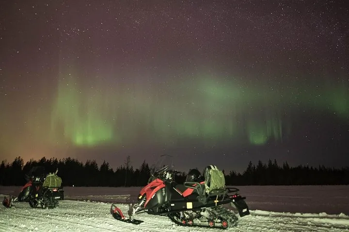 Two red snowmobiles parked on snow under a faint green aurora and starry sky