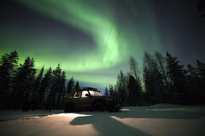 A 4x4 vehicle with its lights on parked in snow under a green aurora