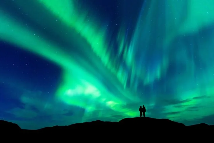 Two people silhouetted on a dark ridge beneath a wide sweep of green aurora